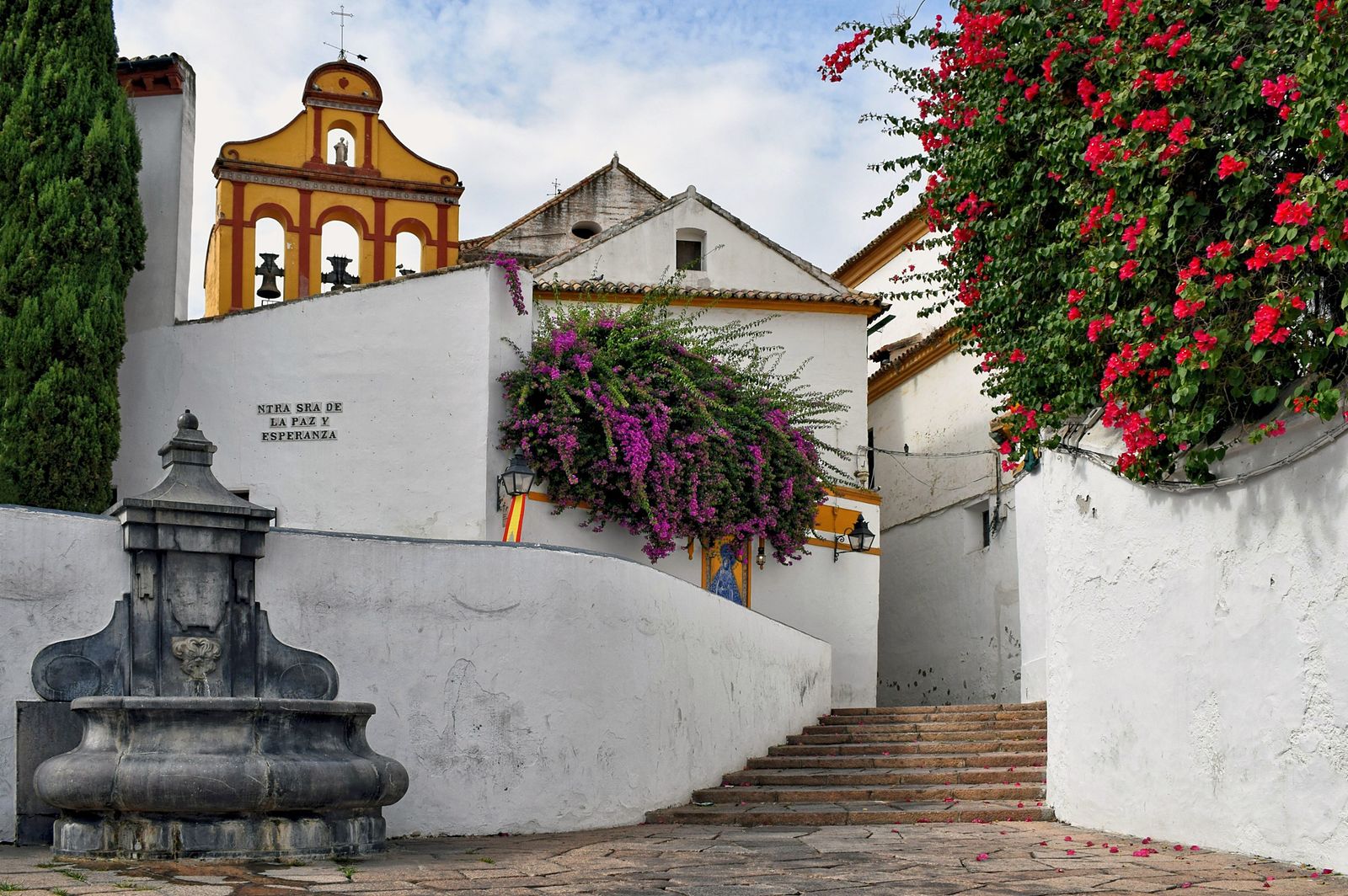 A corner of Córdoba with a fountain, a yellow bell gable and bougainvillea blooming over whitewashed walls.