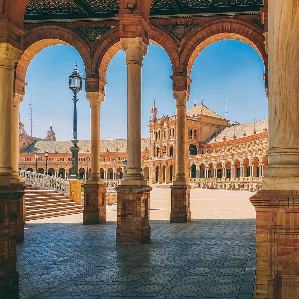 Brick and marble arches frame the Plaza de España in Seville under a blue sky.